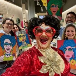 Drag Queen ‘The Fabulous Wonder Mama' dressed in red and smiling for the camera whilst people hold portraits of her in the background